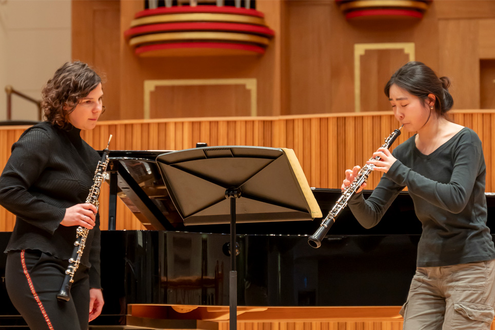 A female student playing the oboe, with a female professor, holding an oboe, observing how the student is playing on a stage, with a piano and wooden panelling behind them.
